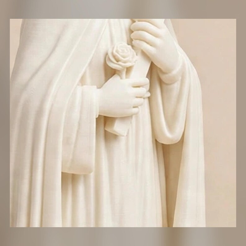 May include: A cream-colored statue featuring a figure draped in flowing, pleated robes. The figure holds a single sculpted rose in one hand and a rectangular object in the other, with both hands positioned centrally against the draped fabric.