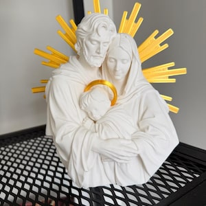 May include: A white matte sculpture of a family of three figures in a close embrace. The figures feature flowing robes and serene expressions. A bright yellow sunburst halo is positioned behind the heads, and a yellow circular halo surrounds the child.