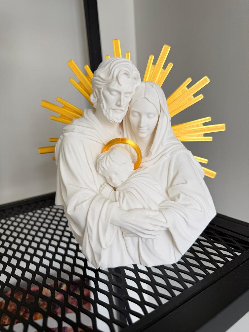 May include: A white matte sculpture of a family of three figures in a close embrace. The figures feature flowing robes and serene expressions. A bright yellow sunburst halo is positioned behind the heads, and a yellow circular halo surrounds the child.