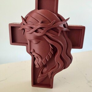 May include: A dark reddish-brown wall cross featuring a three-dimensional relief sculpture of the head of Jesus wearing a crown of thorns. The figure is depicted in profile with long, flowing hair and a beard, set against a simple cross background.