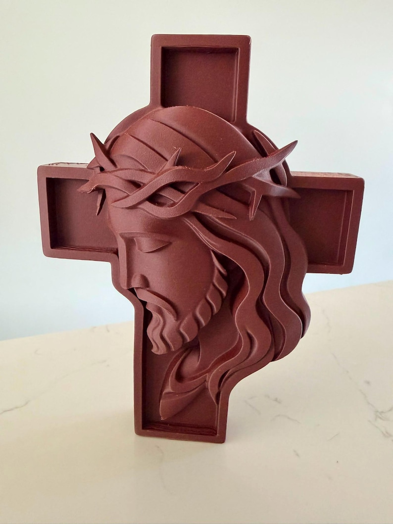 May include: A dark reddish-brown wall cross featuring a three-dimensional relief sculpture of the head of Jesus wearing a crown of thorns. The figure is depicted in profile with long, flowing hair and a beard, set against a simple cross background.