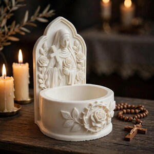 May include: An off-white holy water font featuring a relief sculpture of a nun holding a cross and a rose. The basin is decorated with a carved rose and vine motif. The piece is crafted with a pointed arch backplate and a circular bowl base.