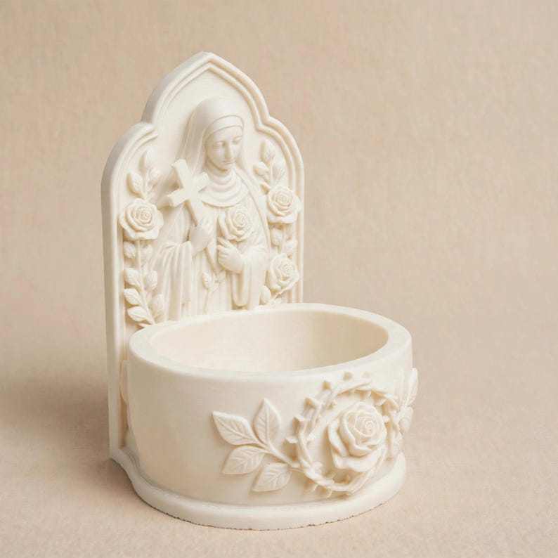 May include: An off-white wall-mounted holy water font featuring a sculpted relief of a figure holding a cross and a rose. The basin is decorated with a carved rose and leafy vine motif, while the back panel displays a pointed arch design.