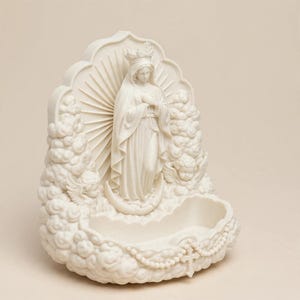 May include: A cream-colored wall-mounted holy water font featuring a central figure of the Virgin Mary wearing a crown and standing on a crescent moon. The figure is surrounded by sculpted clouds, two cherubs, and a rosary with a cross at the base.