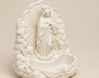 May include: A cream-colored wall-mounted holy water font featuring a central figure of the Virgin Mary wearing a crown and standing on a crescent moon. The figure is surrounded by sculpted clouds, two cherubs, and a rosary with a cross at the base.