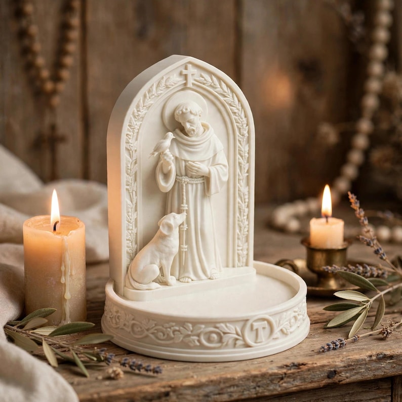 May include: An off-white resin sculpture featuring a robed figure standing under a gothic arch, holding a small bird with a dog sitting at their feet. The base is circular with decorative floral carvings and a prominent letter T embossed on the front.