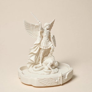 May include: A cream-colored statue of an archangel with large wings, wearing armor and holding a sword raised high in one hand and a shield in the other, standing over a fallen figure on a circular base decorated with carved wings and a sword.