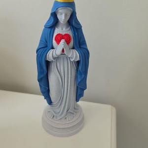 Puede incluir: Una figura de pie que viste una capa azul con capucha y una corona amarilla. Las manos est&aacute;n juntas frente a un emblema rojo en forma de coraz&oacute;n. La base, la t&uacute;nica y la cabeza son de color gris claro, sobre un pedestal circular.