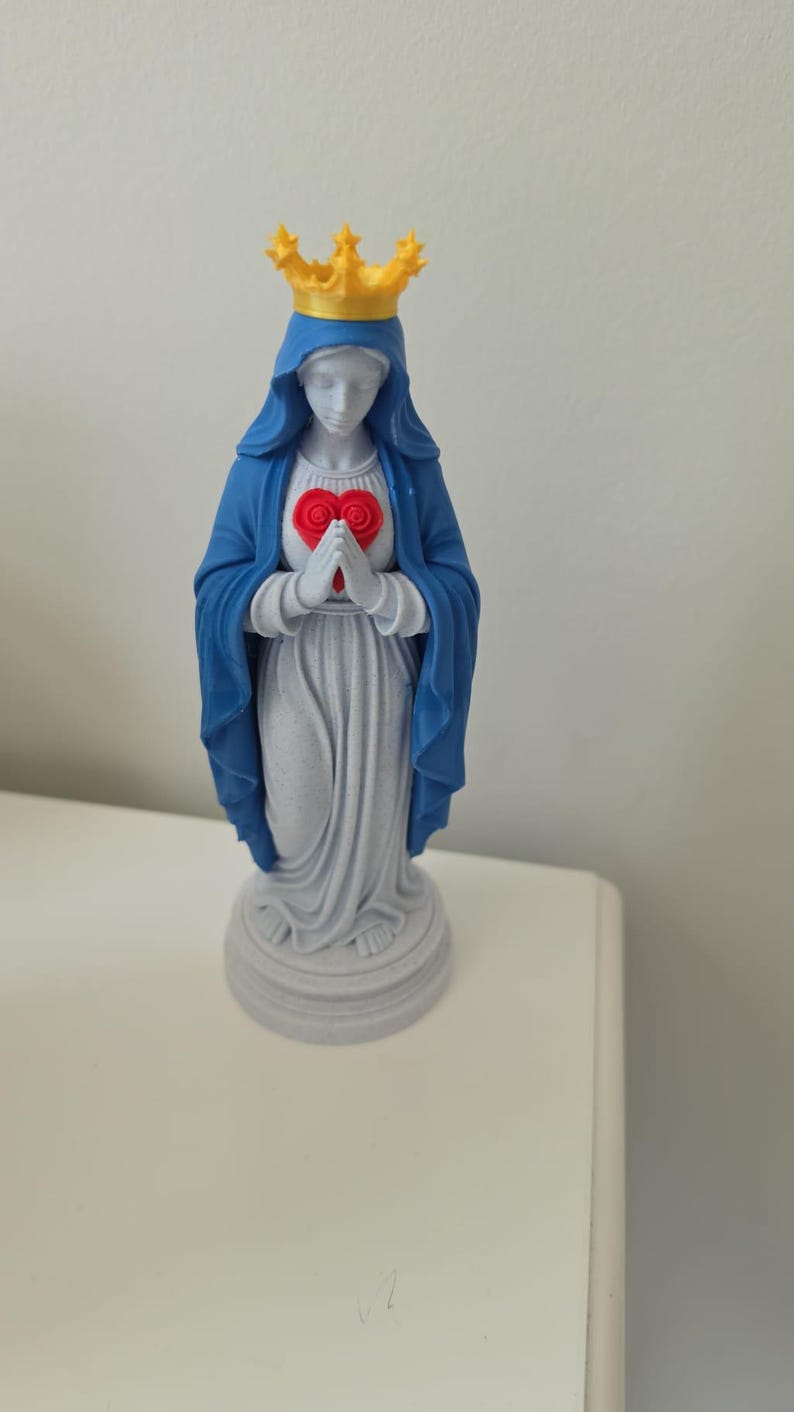 Puede incluir: Una figura de pie que viste una capa azul con capucha y una corona amarilla. Las manos est&aacute;n juntas frente a un emblema rojo en forma de coraz&oacute;n. La base, la t&uacute;nica y la cabeza son de color gris claro, sobre un pedestal circular.