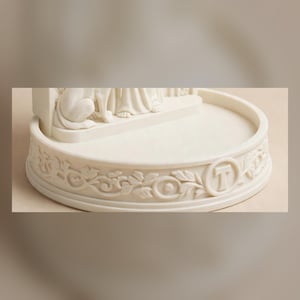 May include: A cream-colored circular base for a figurine featuring an intricate relief of carved leaves and vines wrapping around the perimeter. The front of the base displays a prominent capital letter T centered within a circular frame.