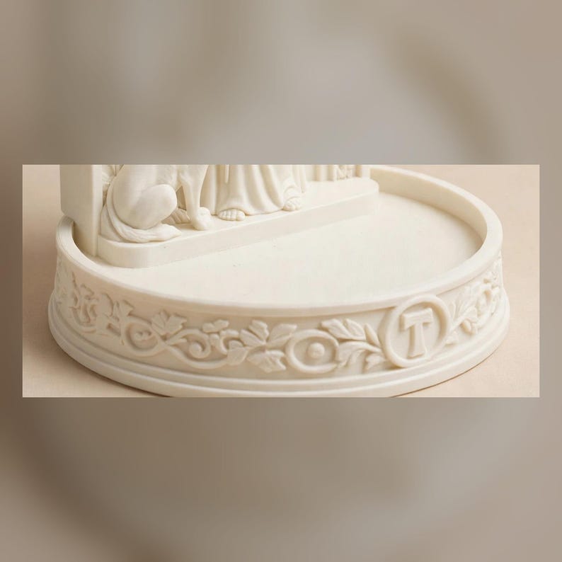 May include: A cream-colored circular base for a figurine featuring an intricate relief of carved leaves and vines wrapping around the perimeter. The front of the base displays a prominent capital letter T centered within a circular frame.