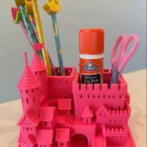 K&ouml;nnte beinhalten: Ein leuchtend pinker Schreibtisch-Organizer aus Kunststoff in Form einer Miniaturburg mit mehreren F&auml;chern. Er enth&auml;lt mehrere bunte Bleistifte mit fruchtf&ouml;rmigen Radiergummis, einen Elmer's Allesklebestift und eine hellrosa Schere.