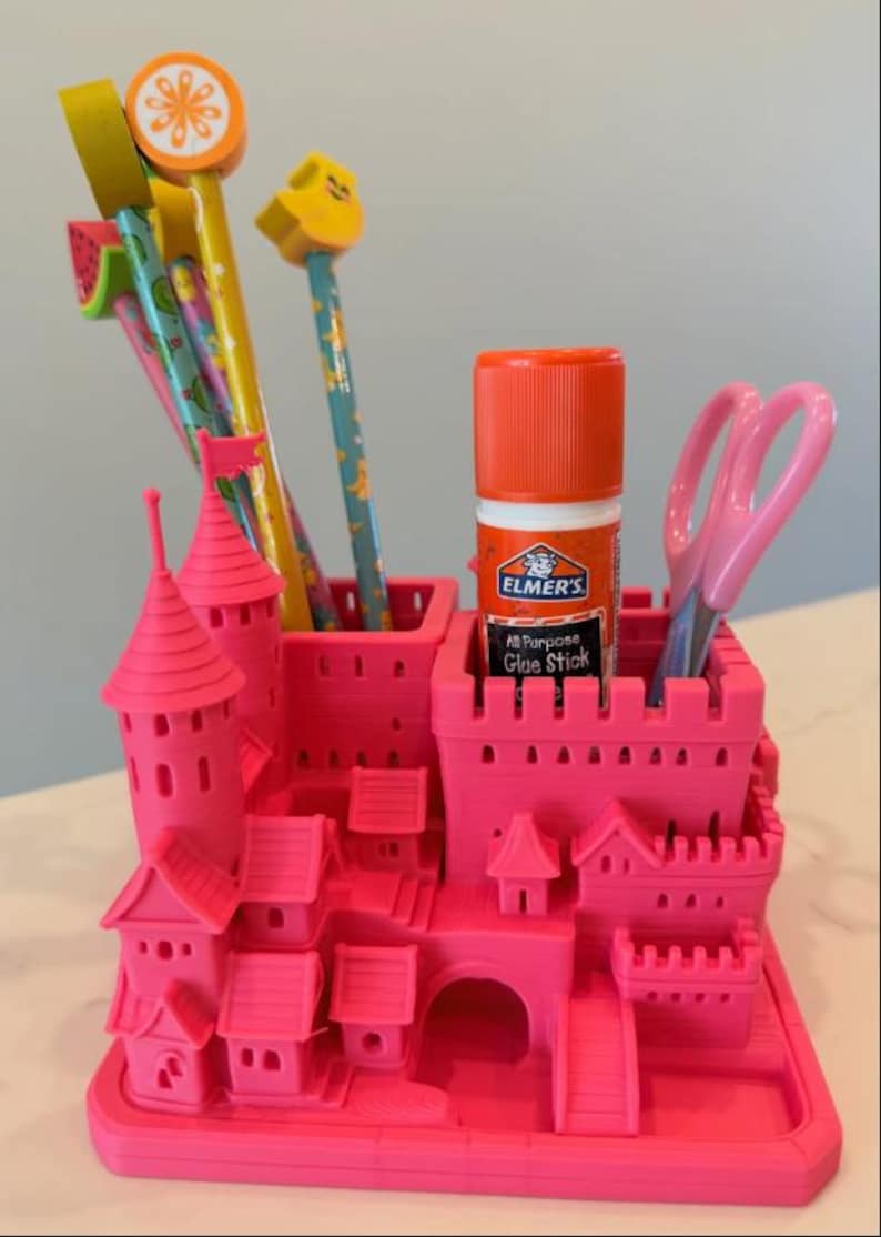 K&ouml;nnte beinhalten: Ein leuchtend pinker Schreibtisch-Organizer aus Kunststoff in Form einer Miniaturburg mit mehreren F&auml;chern. Er enth&auml;lt mehrere bunte Bleistifte mit fruchtf&ouml;rmigen Radiergummis, einen Elmer's Allesklebestift und eine hellrosa Schere.