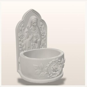 May include: A white wall-mounted holy water font featuring a relief sculpture of a figure in a habit holding a cross and roses. The basin is circular with a relief of a rose surrounded by a crown of thorns on the front exterior surface.