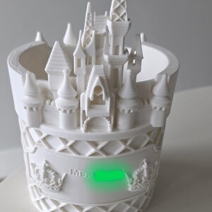 Pu&ograve; includere: Un vaso cilindrico bianco con un design a castello stampato in 3D sul bordo. La parete esterna presenta un motivo a reticolo, due corone in rilievo e il testo "Ms." seguito da un'area verde oscurata. La struttura &egrave; di colore bianco opaco.