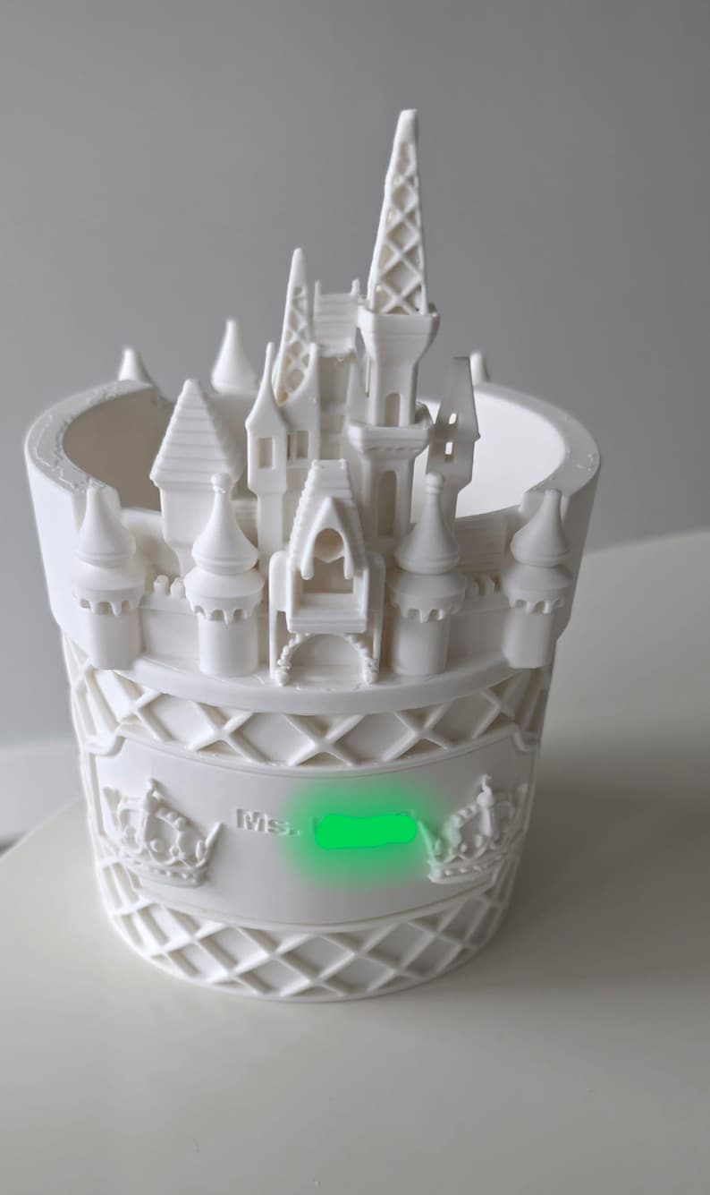 Pu&ograve; includere: Un vaso cilindrico bianco con un design a castello stampato in 3D sul bordo. La parete esterna presenta un motivo a reticolo, due corone in rilievo e il testo "Ms." seguito da un'area verde oscurata. La struttura &egrave; di colore bianco opaco.