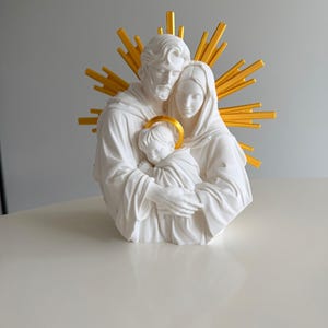 May include: A white sculpture of a family of three figures embracing, featuring a bearded adult, a woman, and a child. The child has a gold halo, and a large, radiating gold sunburst design is positioned behind the figures' heads.