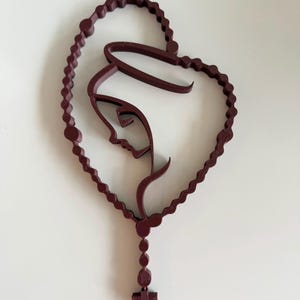 May include: A dark maroon, three-dimensional wall art piece featuring a continuous line drawing of a profile of the Virgin Mary enclosed within a heart-shaped rosary. The design includes a dangling section with beads and a small cross at the bottom.