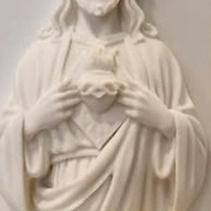 May include: A white wall plaque featuring a relief sculpture of a figure with long hair and a beard, wearing draped robes. The figure holds a heart shape at the centre of the chest with both hands, with flames depicted at the top of the heart.