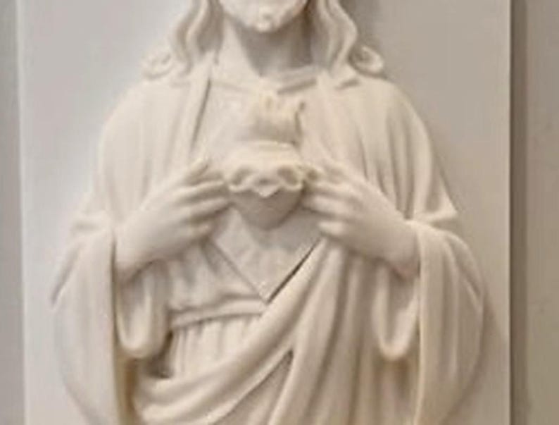 May include: A white wall plaque featuring a relief sculpture of a figure with long hair and a beard, wearing draped robes. The figure holds a heart shape at the centre of the chest with both hands, with flames depicted at the top of the heart.