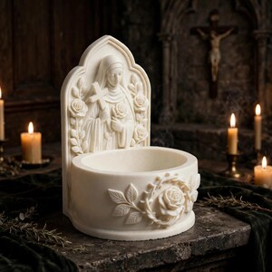May include: A cream-colored holy water font featuring a sculpted figure of a nun holding a cross and a rose. The back panel is arched with floral carvings, while the circular basin is decorated with a prominent rose and leaf relief design.