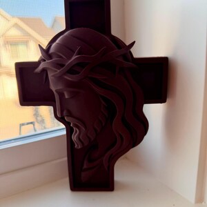 May include: A dark maroon wall-mounted cross featuring a three-dimensional relief sculpture of the face of Jesus wearing a crown of thorns. The sculpture is layered, showing the profile of the face with long hair and a beard against a cross.