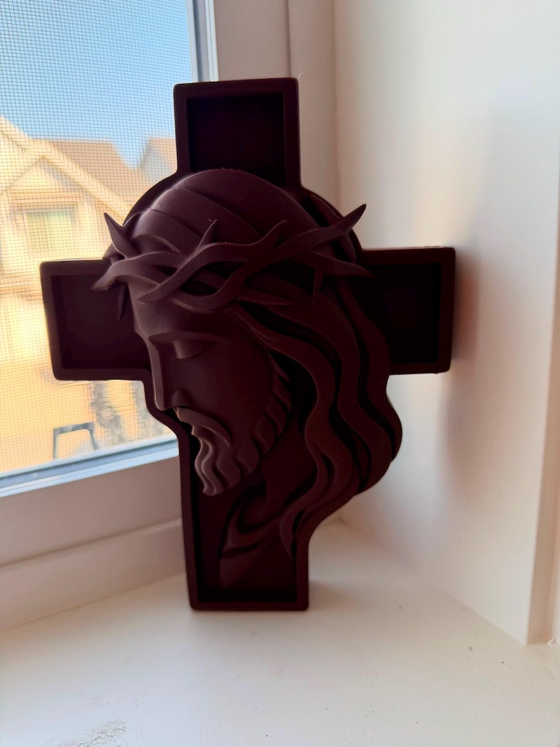 May include: A dark maroon wall-mounted cross featuring a three-dimensional relief sculpture of the face of Jesus wearing a crown of thorns. The sculpture is layered, showing the profile of the face with long hair and a beard against a cross.