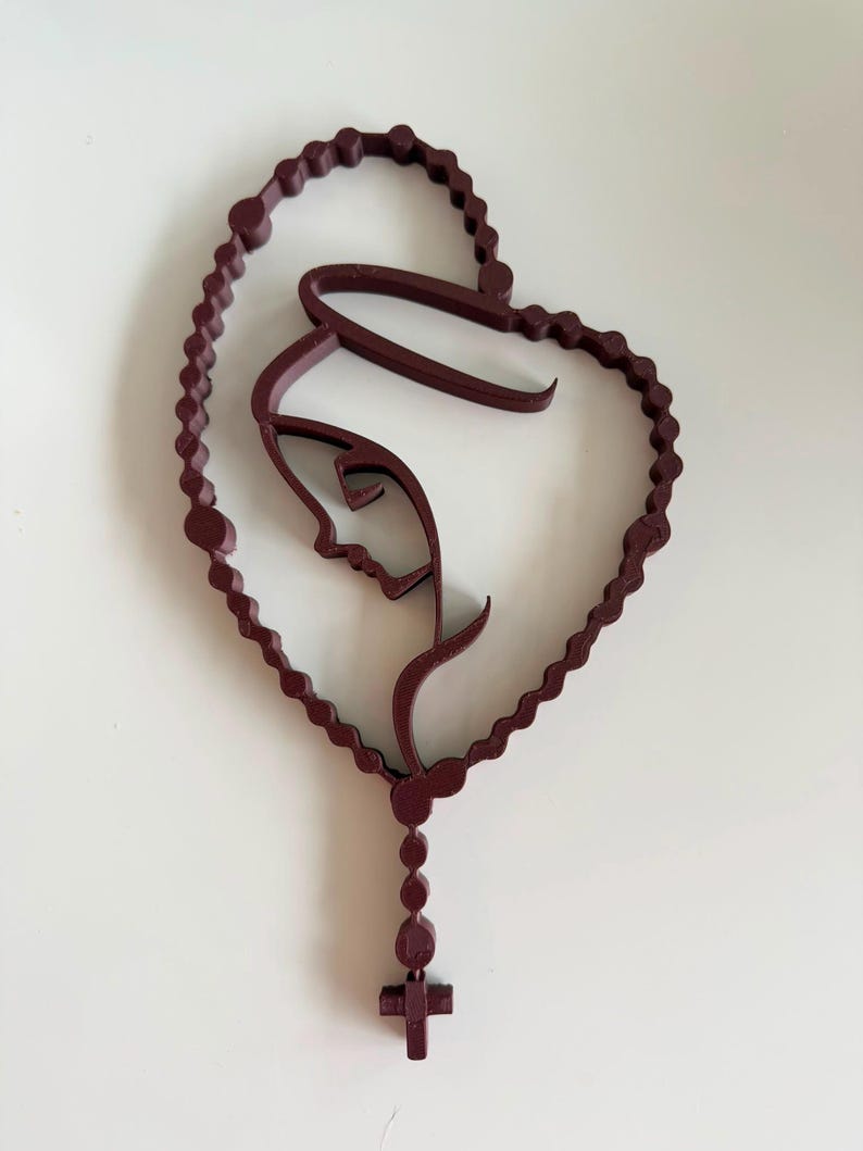 May include: A dark maroon, three-dimensional wall art piece featuring a continuous line drawing of a profile of the Virgin Mary enclosed within a heart-shaped rosary. The design includes a dangling section with beads and a small cross at the bottom.