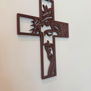 Puede incluir: Una cruz de pared de color marr&oacute;n oscuro con un dise&ntilde;o recortado de una figura que lleva una corona de espinas. La silueta muestra la cabeza y las manos de la figura integradas en las barras verticales y horizontales de la estructura.