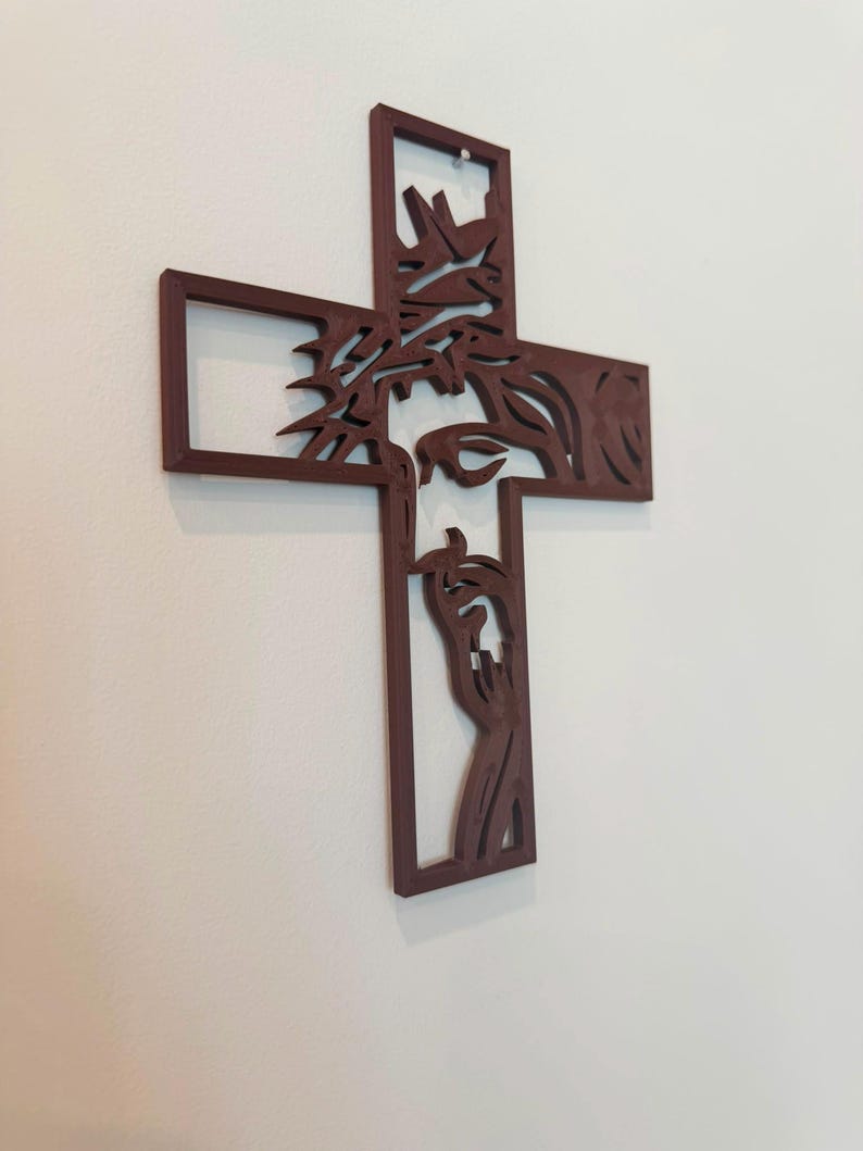 Puede incluir: Una cruz de pared de color marr&oacute;n oscuro con un dise&ntilde;o recortado de una figura que lleva una corona de espinas. La silueta muestra la cabeza y las manos de la figura integradas en las barras verticales y horizontales de la estructura.
