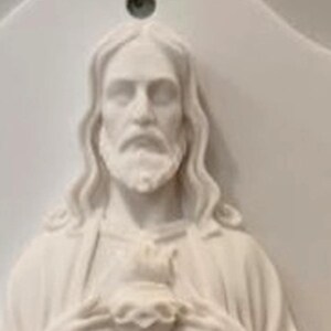 May include: A white wall plaque featuring a relief sculpture of a bearded figure with long hair, holding a heart at the chest. The plaque has a smooth, matte finish and includes a small circular hole at the top centre for hanging on a wall.