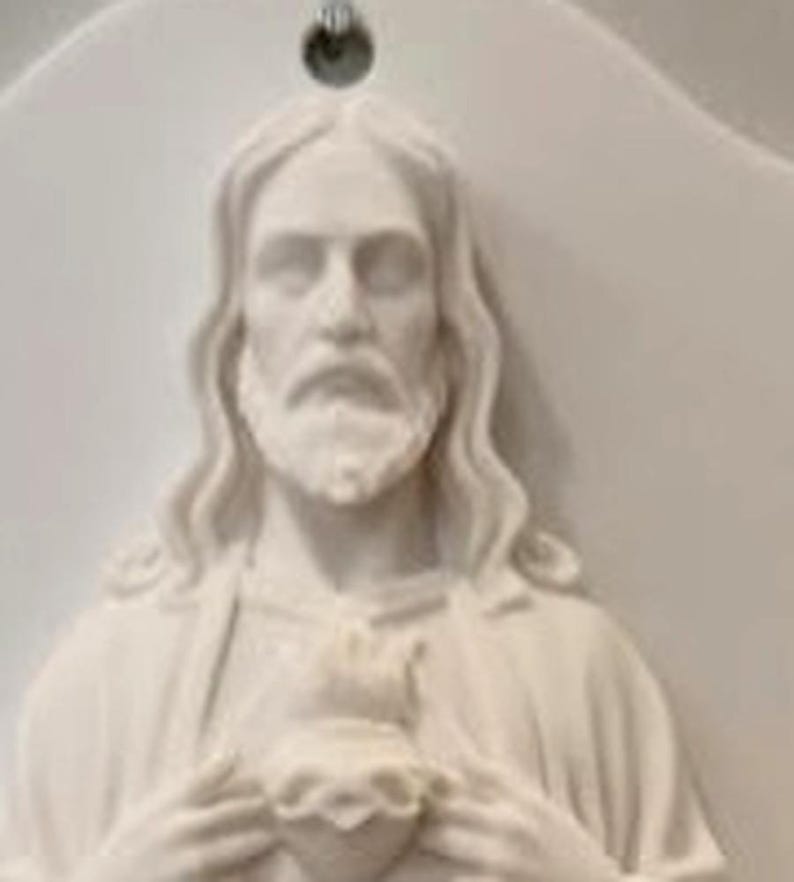 May include: A white wall plaque featuring a relief sculpture of a bearded figure with long hair, holding a heart at the chest. The plaque has a smooth, matte finish and includes a small circular hole at the top centre for hanging on a wall.