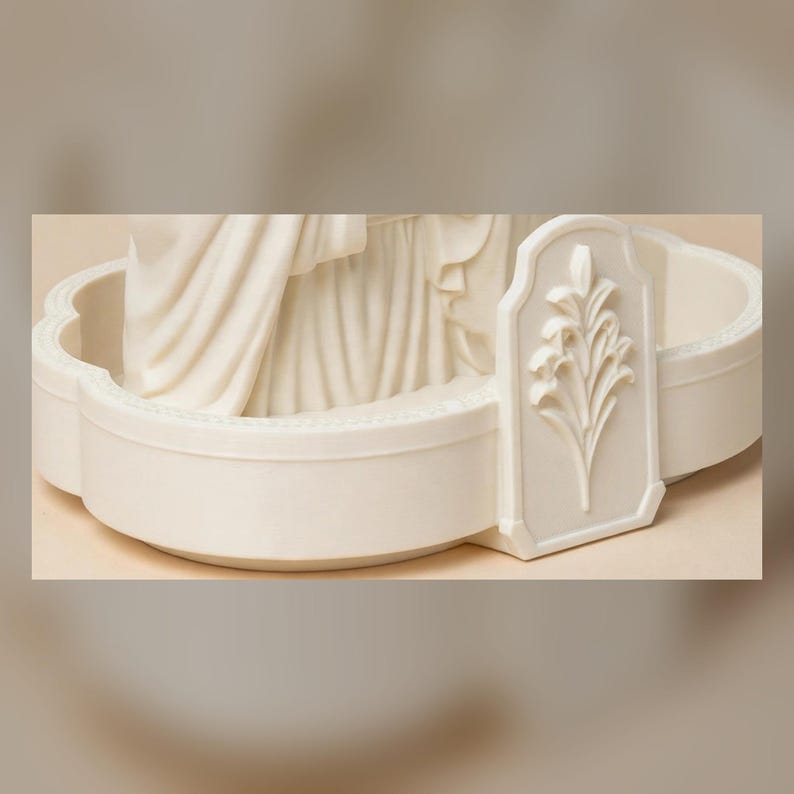 May include: A cream-colored, off-white circular base for a figurine, featuring a scalloped edge and a raised, rectangular plaque on the front. The plaque displays a detailed, embossed relief of a stylized floral plant with multiple leaves.