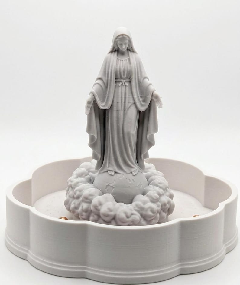 May include: A light gray 3D printed figurine of a standing figure with a draped head covering and long robes, arms outstretched, positioned atop a spherical base surrounded by sculpted clouds, all set within a circular scalloped tray.