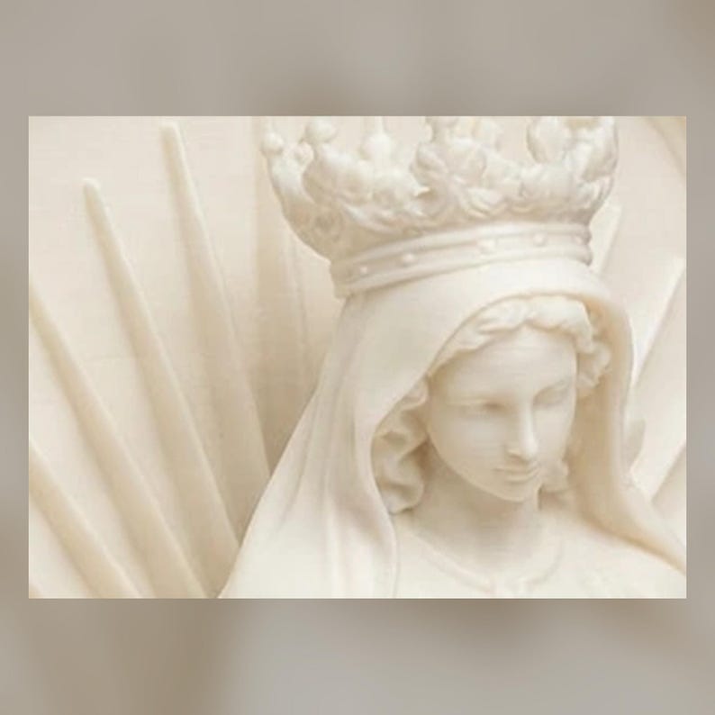 May include: A white marble-like sculpture depicting a figure wearing a crown and a draped head covering. The figure has a serene facial expression, and the background features radiating sunburst rays carved in a relief pattern behind the head.