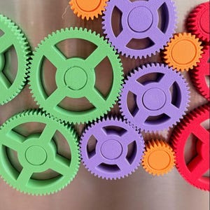 K&ouml;nnte beinhalten: Eine Sammlung von zahnradf&ouml;rmigen Kunststoffmagneten in verschiedenen Gr&ouml;&szlig;en und Farben, darunter Gr&uuml;n, Lila, Orange und Rot. Jedes Zahnrad verf&uuml;gt &uuml;ber eine zentrale Nabe mit vier Speichen und eine texturierte Oberfl&auml;che.