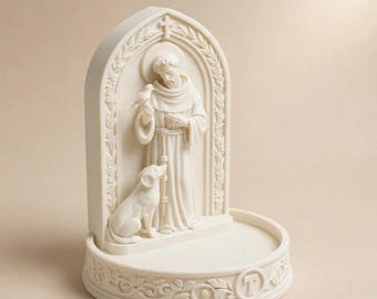May include: An off-white, cream-colored resin sculpture featuring a figure in robes standing within an arched frame. The figure holds a small bird, with a dog sitting at their feet. The base is circular and decorated with floral carvings and a letter T.