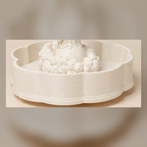 May include: A cream-colored, circular tray with a scalloped edge and a smooth finish. In the center, a sculpted, off-white figure sits atop a spherical base, surrounded by a ring of textured, cloud-like or floral decorative elements.