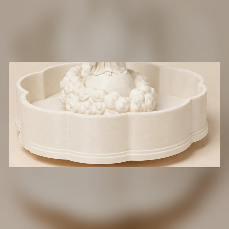 May include: A cream-colored, circular tray with a scalloped edge and a smooth finish. In the center, a sculpted, off-white figure sits atop a spherical base, surrounded by a ring of textured, cloud-like or floral decorative elements.