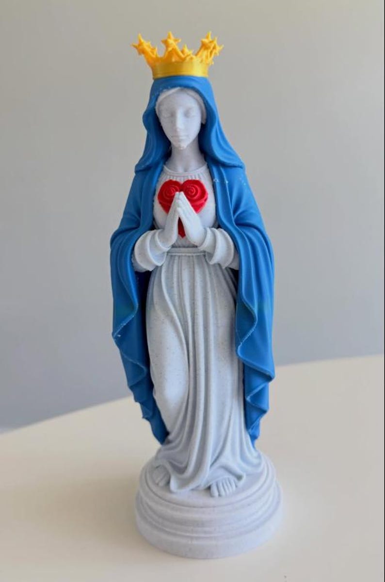 Puede incluir: Una figura blanca que viste un velo azul y una t&uacute;nica larga blanca, sobre una base circular. Las manos est&aacute;n juntas frente a un coraz&oacute;n rojo y lleva una corona amarilla con puntas en forma de estrella sobre la cabeza.