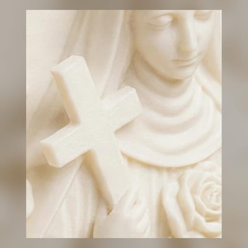 May include: A cream-colored sculpture depicting a figure with a draped head covering, holding a plain cross in one hand and a sculpted rose flower in the other. The surface has a smooth, matte finish with soft contours defining the facial features.