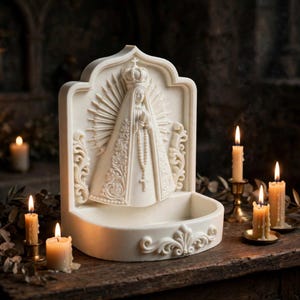 Puede incluir: Una pila de agua bendita de pared en color crema, blanco roto, que presenta una escultura en relieve de la Virgen Mar&iacute;a con corona y rosario, con un halo de rayos detr&aacute;s y adornos de volutas decorativas en los laterales y la base.