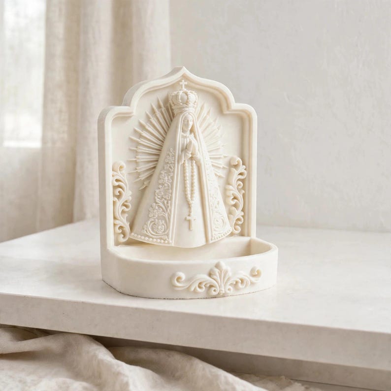 Puede incluir: Una pila de agua bendita de color crema con una escultura en relieve de la Virgen Mar&iacute;a, que lleva una corona y una t&uacute;nica larga con detalles florales. La figura est&aacute; enmarcada por un halo radiante y volutas, con un cuenco en la base.