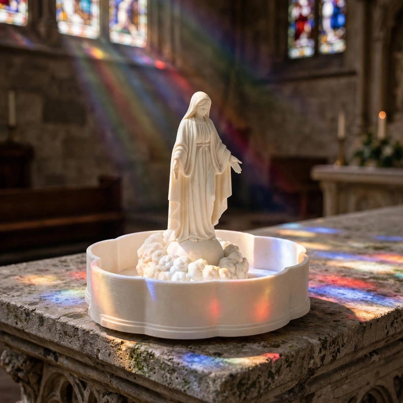 May include: A white statue of the Virgin Mary standing on a cloud base, positioned within a circular, scalloped-edge tray. The figure wears a long, flowing robe with arms extended downward, crafted from a smooth, matte-finish material.