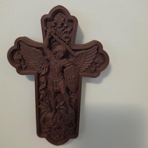 May include: A dark brown wall-mounted cross featuring a detailed relief carving of an angel with wings, holding a sword and a shield, standing over a dragon. The cross has an ornate, scalloped edge design with decorative floral motifs at the top.