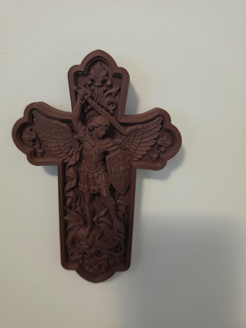 May include: A dark brown wall-mounted cross featuring a detailed relief carving of an angel with wings, holding a sword and a shield, standing over a dragon. The cross has an ornate, scalloped edge design with decorative floral motifs at the top.