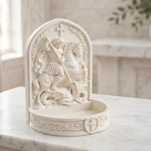 K&ouml;nnte beinhalten: Ein cremefarbener, bogenf&ouml;rmiger Weihwasserbecken zur Wandmontage mit einem Relief von St. Georg zu Pferd, der einen Drachen besiegt. Der Sockel enth&auml;lt ein kleines Becken f&uuml;r Wasser, verziert mit floralen Schnitzereien und einem Wappenschild.