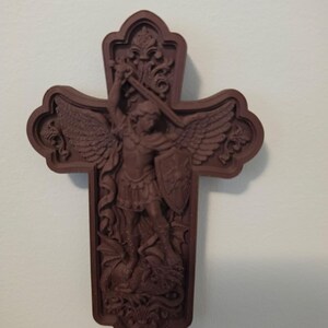 May include: A dark brown wall-mounted cross featuring a relief carving of Saint Michael the Archangel with wings, holding a sword and shield while standing over a dragon. The cross has an ornate, decorative border with intricate scrollwork details.