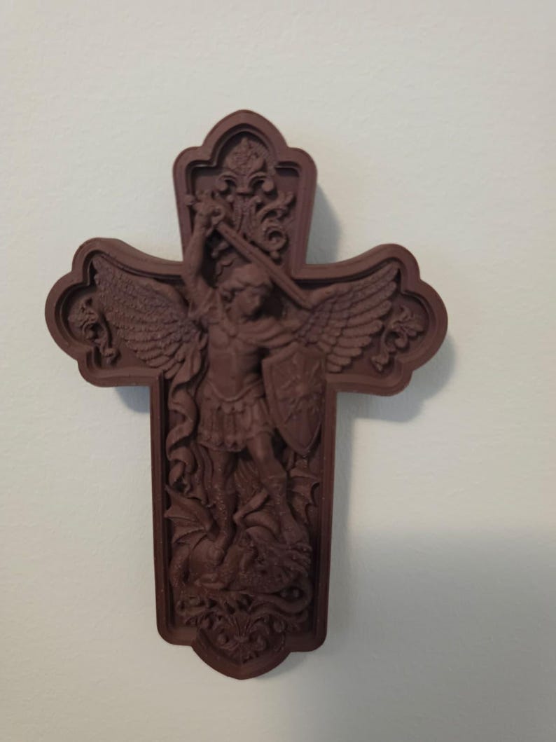 May include: A dark brown wall-mounted cross featuring a relief carving of Saint Michael the Archangel with wings, holding a sword and shield while standing over a dragon. The cross has an ornate, decorative border with intricate scrollwork details.