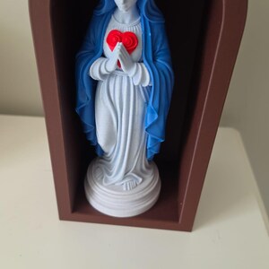 Puede incluir: Una figura blanca que viste una capa azul y una corona dorada, sosteniendo un coraz&oacute;n rojo con ambas manos. La figura est&aacute; colocada dentro de un marco marr&oacute;n arqueado de frente abierto sobre una base circular blanca.